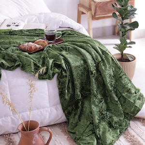 Peut inclure: Une couverture en velours vert olive avec un motif floral, drapée sur un lit blanc. Un plateau en bois contient une tasse de café et des pâtisseries. La scène comprend un vase en terre cuite et une plante en pot, créant une atmosphère confortable.