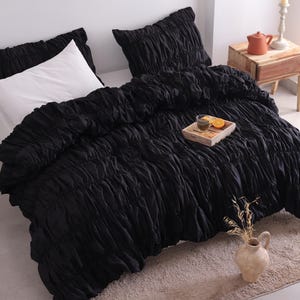 May include: A black ruched duvet cover and pillow set on a bed. A white pillow is visible. A small wooden tray with a drink and orange slices sits on the duvet. A small vase with dried flowers is in the foreground.