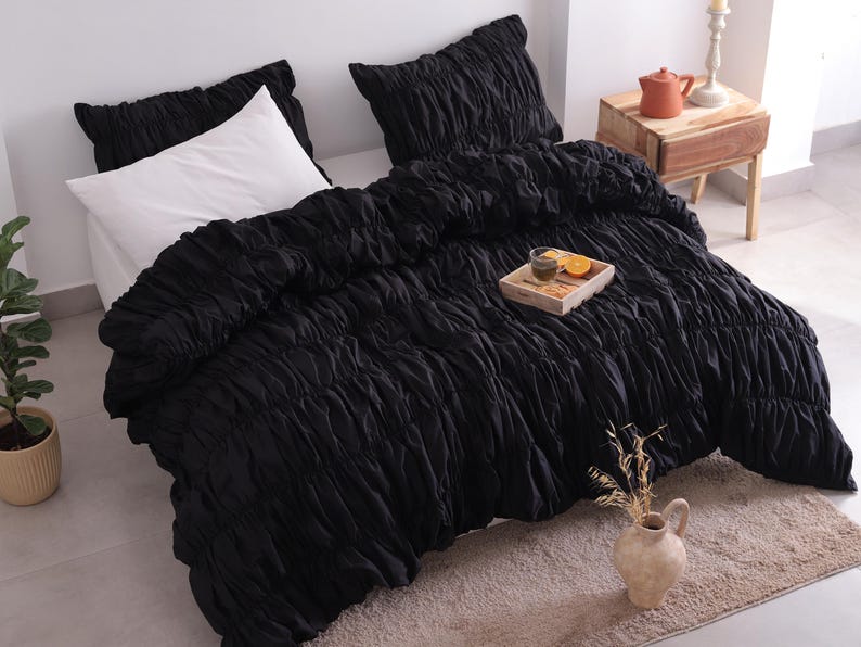May include: A black ruched duvet cover and pillow set on a bed. A white pillow is visible. A small wooden tray with a drink and orange slices sits on the duvet. A small vase with dried flowers is in the foreground.