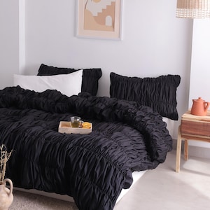 May include: A bed dressed with black ruched bedding, two black ruched pillows, and a white pillow. A wooden tray with a drink and fruit sits on the bed. A framed print hangs on the wall.