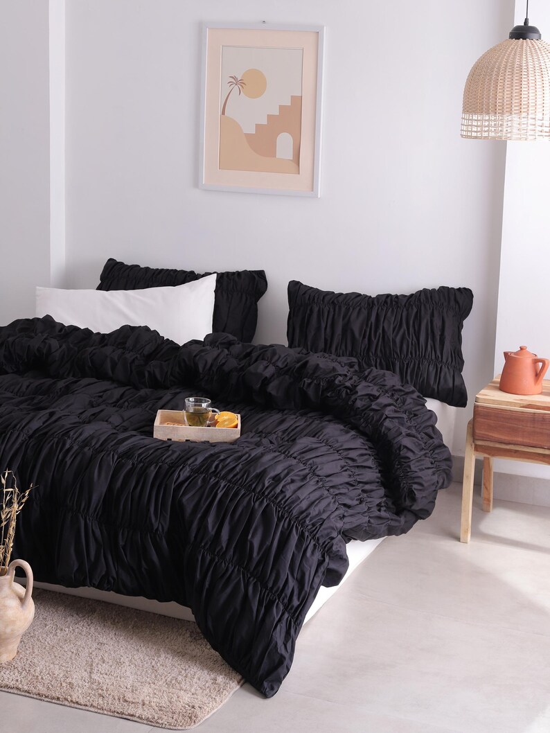 May include: A bed dressed with black ruched bedding, two black ruched pillows, and a white pillow. A wooden tray with a drink and fruit sits on the bed. A framed print hangs on the wall.