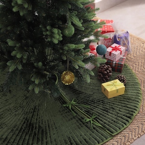 Puede incluir: Un &aacute;rbol de Navidad decorado con adornos y regalos. Una falda de &aacute;rbol plisada de color verde oscuro con ribete y lazos verdes rodea la base del &aacute;rbol. El &aacute;rbol se encuentra sobre una alfombra tejida de color natural.