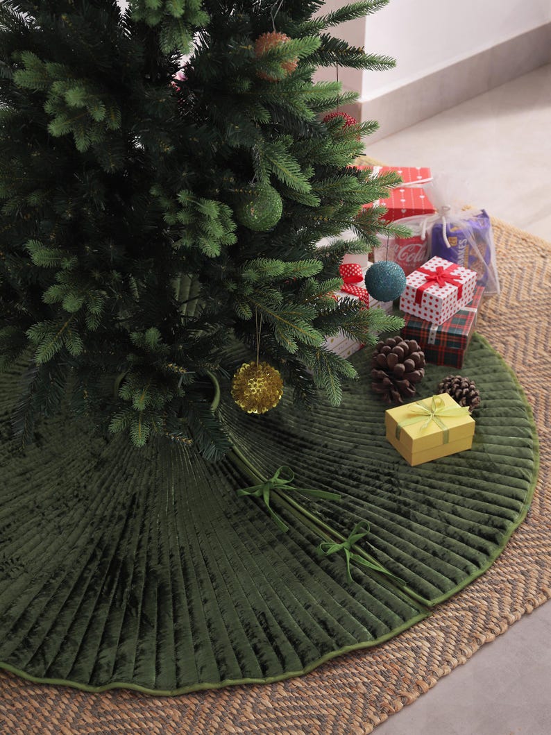 Puede incluir: Un &aacute;rbol de Navidad decorado con adornos y regalos. Una falda de &aacute;rbol plisada de color verde oscuro con ribete y lazos verdes rodea la base del &aacute;rbol. El &aacute;rbol se encuentra sobre una alfombra tejida de color natural.