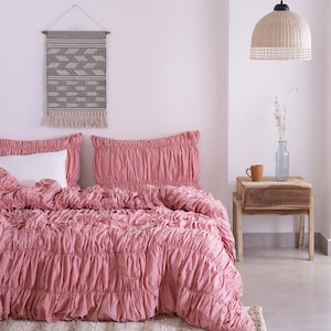 May include: A bedroom scene featuring a bed with a pink ruched comforter and pillowcases. A white pillow is visible. A woven wall hanging with a geometric pattern hangs above the bed. A woven pendant light and a wooden nightstand with a mug and vase are also present.