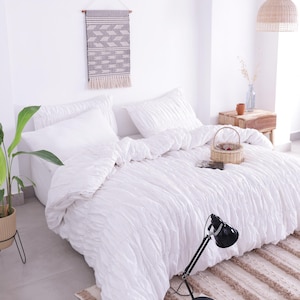 May include: A bright, airy bedroom scene featuring a white bed with a textured comforter and pillows. A small woven basket sits on the bed. A black desk lamp and a patterned rug add contrast. A woven wall hanging and a potted plant complete the decor.