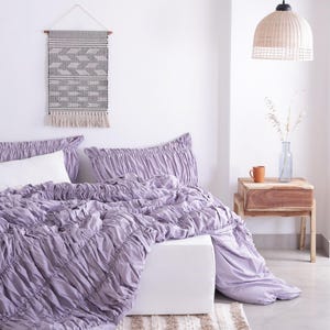 Puede incluir: Una escena de dormitorio con una colcha fruncida lavanda y fundas de almohada a juego. Se ve una almohada blanca. Un tapiz tejido con un patr&oacute;n geom&eacute;trico cuelga sobre la cama. Una mesita de noche de madera con una taza y un jarr&oacute;n.