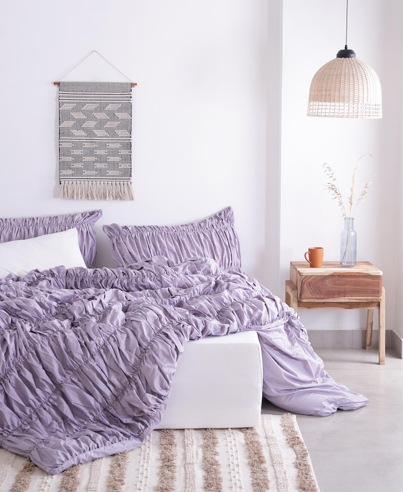 Puede incluir: Una escena de dormitorio con una colcha fruncida lavanda y fundas de almohada a juego. Se ve una almohada blanca. Un tapiz tejido con un patr&oacute;n geom&eacute;trico cuelga sobre la cama. Una mesita de noche de madera con una taza y un jarr&oacute;n.