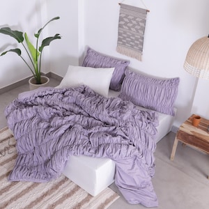 Puede incluir: Una cama con un edred&oacute;n fruncido de color lavanda y fundas de almohada a juego. Una almohada blanca se coloca detr&aacute;s de las almohadas lavanda. Una planta en maceta, una peque&ntilde;a mesa de madera y una l&aacute;mpara colgante tejida tambi&eacute;n est&aacute;n en la habitaci&oacute;n.