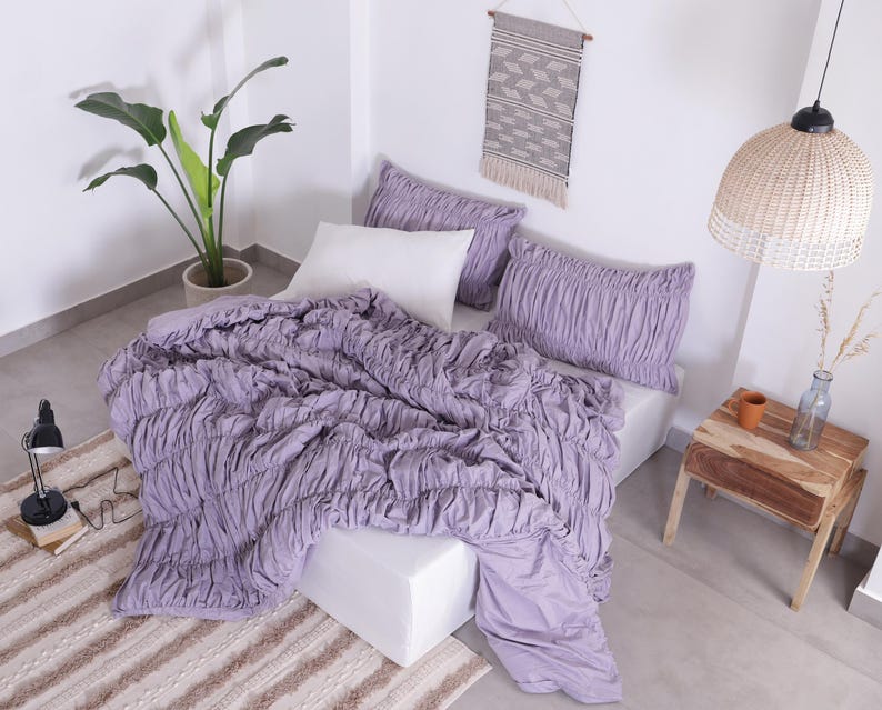 Puede incluir: Una cama con un edred&oacute;n fruncido de color lavanda y fundas de almohada a juego. Una almohada blanca se coloca detr&aacute;s de las almohadas lavanda. Una planta en maceta, una peque&ntilde;a mesa de madera y una l&aacute;mpara colgante tejida tambi&eacute;n est&aacute;n en la habitaci&oacute;n.