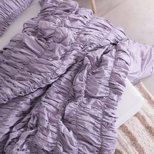 Puede incluir: Un juego de cama de color lavanda con un dise&ntilde;o fruncido. El juego incluye un edred&oacute;n y una funda de almohada. La ropa de cama est&aacute; dispuesta sobre una superficie blanca, con una alfombra de tonos neutros visible. La tela tiene un aspecto suave y texturizado.