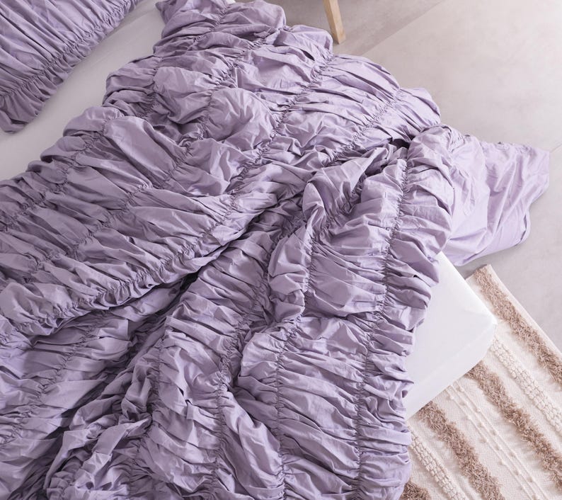 Puede incluir: Un juego de cama de color lavanda con un dise&ntilde;o fruncido. El juego incluye un edred&oacute;n y una funda de almohada. La ropa de cama est&aacute; dispuesta sobre una superficie blanca, con una alfombra de tonos neutros visible. La tela tiene un aspecto suave y texturizado.