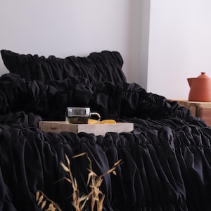 May include: Black bedding with a ruffled design. A wooden tray holds a glass of tea and a slice of orange. A terracotta teapot is in the background. Dried plants are in the foreground.