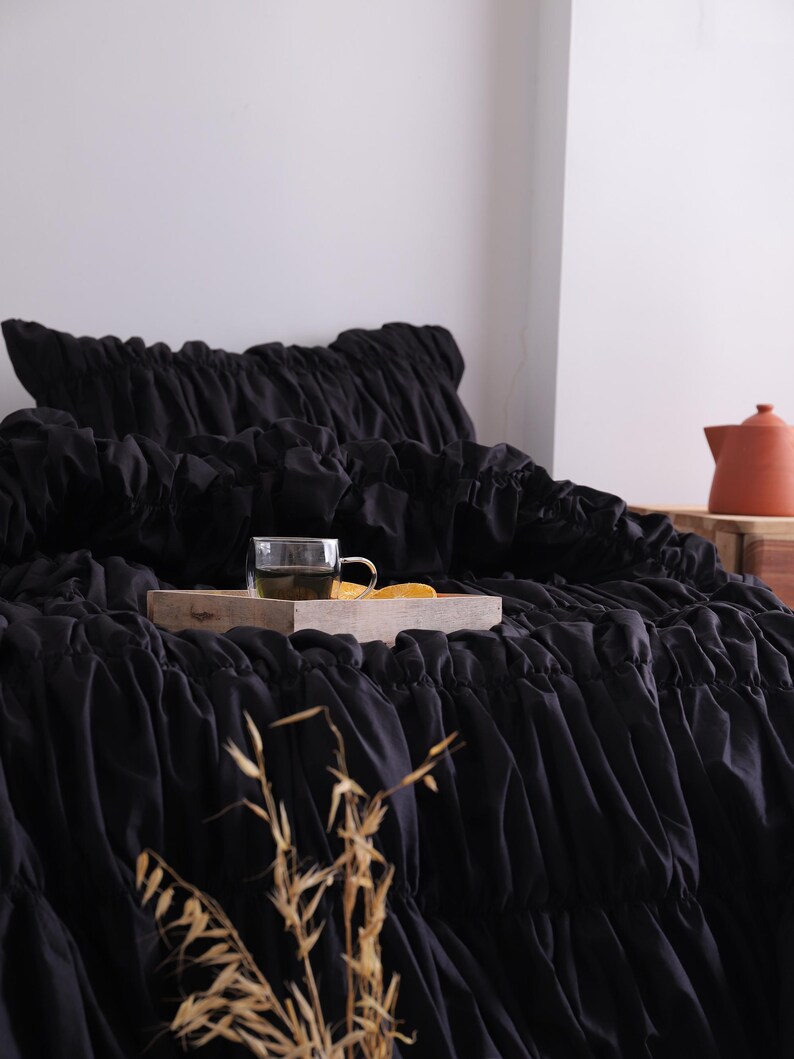 May include: Black bedding with a ruffled design. A wooden tray holds a glass of tea and a slice of orange. A terracotta teapot is in the background. Dried plants are in the foreground.