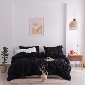 May include: A black bedspread and pillow set with a textured design. The bed is made up with white pillows and a decorative tray. A framed print hangs on the white wall above the bed. A beige rug is at the foot of the bed.
