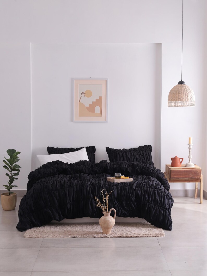 May include: A black bedspread and pillow set with a textured design. The bed is made up with white pillows and a decorative tray. A framed print hangs on the white wall above the bed. A beige rug is at the foot of the bed.