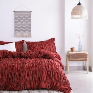 Peut inclure: Une chambre à coucher avec une housse de couette et un ensemble d'oreillers froncés de couleur rouille. Un oreiller blanc est visible. Une tenture murale tissée avec un motif géométrique est suspendue au-dessus du lit. Une suspension tressée et une table de chevet en bois complètent la scène.