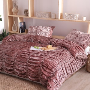 Puede incluir: Un juego de funda nórdica de terciopelo rosa empolvado con una superficie muy fruncida y texturizada. La ropa de cama incluye un par de fundas de almohada rectangulares a juego con el mismo diseño de tela recogida, creando un aspecto suave.