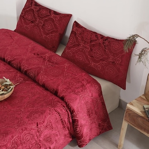 May include: A bed with a deep red paisley patterned duvet cover and two matching pillowcases. A small wooden bowl with dried fruit and greenery sits on the bed. A small wooden side table holds a glass of wine and a decorative vase.
