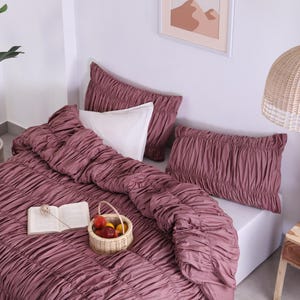 May include: A bed with a mauve ruched comforter and matching pillows. A white pillow is visible. A small basket of fruit and an open book rest on the comforter. A woven pendant light hangs above a wooden side table.