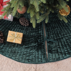 May include: A dark green velvet Christmas tree skirt with a pleated design. The skirt is under a decorated Christmas tree with ornaments and a gift box. Pine cones and a small gift box are on the skirt.