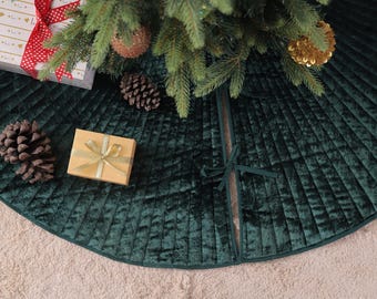 Puede incluir: Una falda de árbol de Navidad de terciopelo verde oscuro con un diseño plisado. La falda está debajo de un árbol de Navidad decorado con adornos y una caja de regalo. Piñas y una pequeña caja de regalo están en la falda.