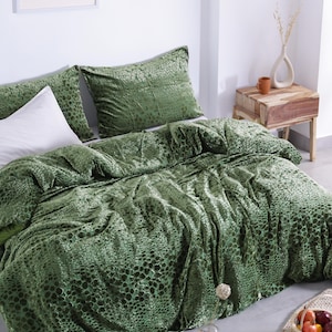 Puede incluir: Una cama está hecha con un edredón verde estampado y fundas de almohada a juego. La ropa de cama tiene un aspecto texturizado, similar al terciopelo. También se ve una almohada blanca. La habitación tiene una paleta de colores neutros con una mesita de noche de madera.