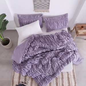 Puede incluir: Una cama est&aacute; hecha con un edred&oacute;n fruncido de color lavanda y dos fundas de almohada a juego. Una almohada blanca se coloca en el centro. La cama est&aacute; sobre una alfombra estampada. Una planta en maceta y una peque&ntilde;a mesa de madera est&aacute;n en el fondo.