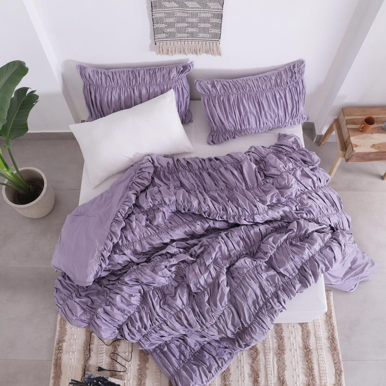 Puede incluir: Una cama est&aacute; hecha con un edred&oacute;n fruncido de color lavanda y dos fundas de almohada a juego. Una almohada blanca se coloca en el centro. La cama est&aacute; sobre una alfombra estampada. Una planta en maceta y una peque&ntilde;a mesa de madera est&aacute;n en el fondo.