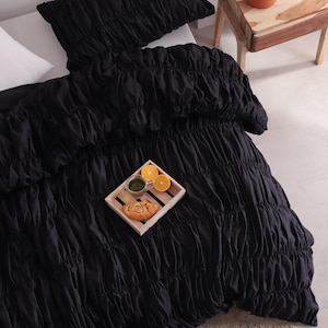 May include: Black ruched bedding set, including a duvet cover and pillow, on a white bed. A wooden tray with food and drinks sits on the duvet. A small wooden bedside table with a terracotta pot and lamp is next to the bed.