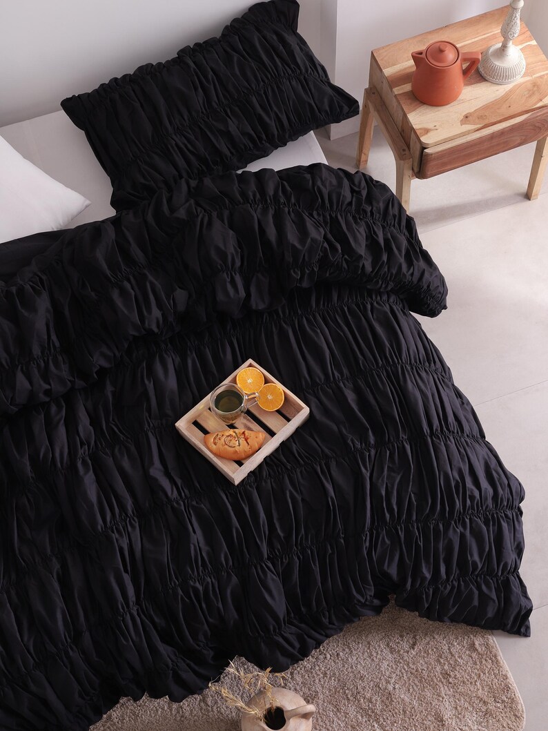 May include: Black ruched bedding set, including a duvet cover and pillow, on a white bed. A wooden tray with food and drinks sits on the duvet. A small wooden bedside table with a terracotta pot and lamp is next to the bed.