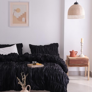 May include: A bedroom scene with a black ruched duvet cover and pillow set. A wooden tray holds a glass and orange slices. A framed print hangs on the wall, and a woven pendant light hangs from the ceiling. A vase with dried plants sits on the floor.