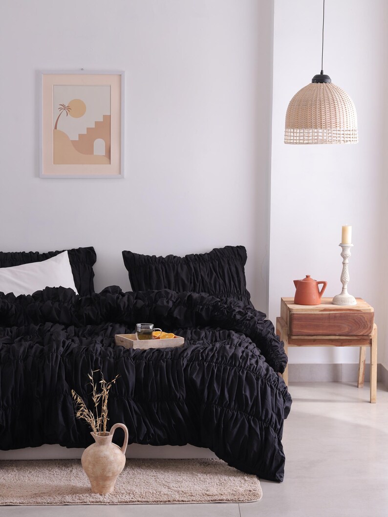 May include: A bedroom scene with a black ruched duvet cover and pillow set. A wooden tray holds a glass and orange slices. A framed print hangs on the wall, and a woven pendant light hangs from the ceiling. A vase with dried plants sits on the floor.