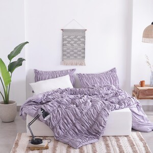 Puede incluir: Dormitorio con una cama con colcha y almohadas lavanda fruncidas. Una almohada blanca est&aacute; sobre la cama. Un tapiz tejido y una planta en maceta completan la decoraci&oacute;n. Una l&aacute;mpara de escritorio negra est&aacute; sobre una alfombra.