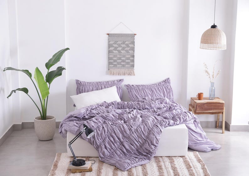 Puede incluir: Dormitorio con una cama con colcha y almohadas lavanda fruncidas. Una almohada blanca est&aacute; sobre la cama. Un tapiz tejido y una planta en maceta completan la decoraci&oacute;n. Una l&aacute;mpara de escritorio negra est&aacute; sobre una alfombra.