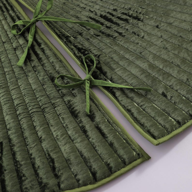 Puede incluir: Primer plano de una tela de terciopelo verde bosque acolchada con costuras de canal verticales. Los bordes est&aacute;n ribeteados con un borde verde a juego. Dos cintas de terciopelo verde est&aacute;n atadas en lazos.