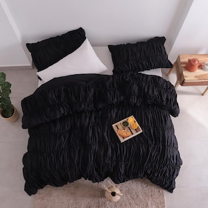 May include: An overhead view of a bed featuring a black ruched duvet cover and two matching pillowcases. A white pillow is visible. A small wooden side table holds a teapot and lamp. A tray with food and drinks sits on the bed.