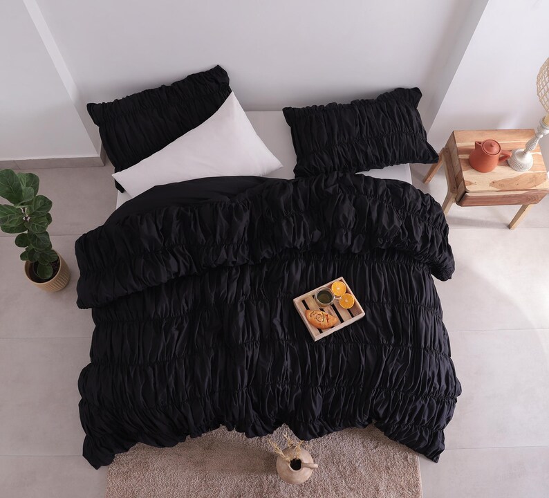 May include: An overhead view of a bed featuring a black ruched duvet cover and two matching pillowcases. A white pillow is visible. A small wooden side table holds a teapot and lamp. A tray with food and drinks sits on the bed.