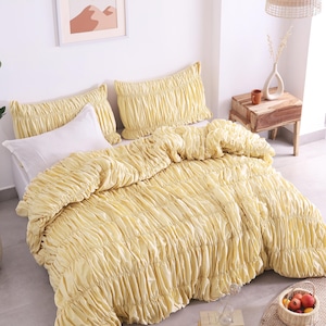May include: A bed set featuring a yellow ruched comforter and matching pillow shams. A white pillow is also visible. The bedding is arranged on a bed, with a framed art print on the wall. A small wooden side table is next to the bed.