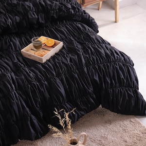 May include: Black ruched bedding on a bed. A wooden tray holds a glass of tea, orange slices, and pastries. A beige rug and a vase with dried plants are in the foreground.