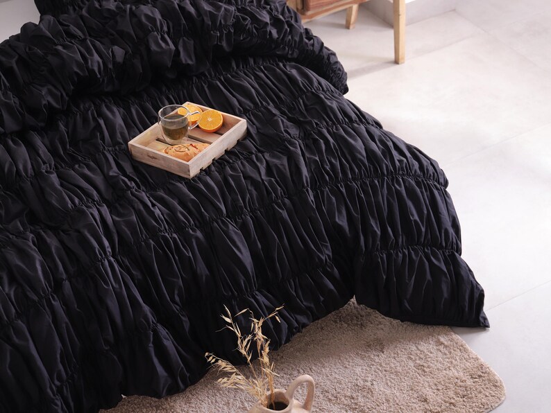 May include: Black ruched bedding on a bed. A wooden tray holds a glass of tea, orange slices, and pastries. A beige rug and a vase with dried plants are in the foreground.