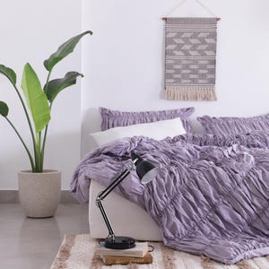 Puede incluir: Una habitaci&oacute;n con una cama cubierta con un edred&oacute;n y almohadas de color lavanda fruncido. Una l&aacute;mpara de escritorio negra descansa sobre libros. A la izquierda hay una gran planta en maceta con hojas verdes y una decoraci&oacute;n colgante de pared sobre la cama.