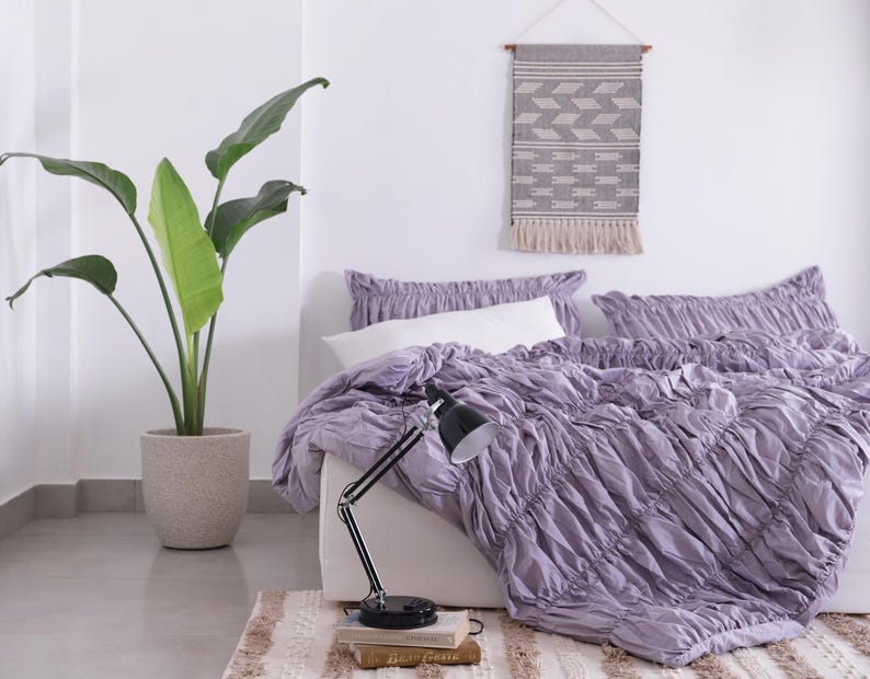 Puede incluir: Una habitaci&oacute;n con una cama cubierta con un edred&oacute;n y almohadas de color lavanda fruncido. Una l&aacute;mpara de escritorio negra descansa sobre libros. A la izquierda hay una gran planta en maceta con hojas verdes y una decoraci&oacute;n colgante de pared sobre la cama.