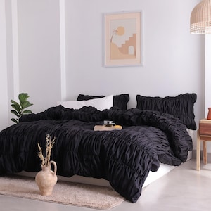 May include: A black ruched duvet cover and pillow set on a bed. The bedding is the focal point, with a neutral-toned rug and a decorative vase with dried stems in the foreground. A framed art piece hangs on the wall.