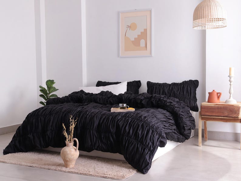May include: A black ruched duvet cover and pillow set on a bed. The bedding is the focal point, with a neutral-toned rug and a decorative vase with dried stems in the foreground. A framed art piece hangs on the wall.