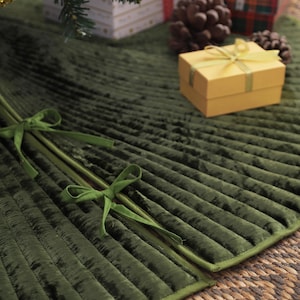 Puede incluir: Una falda de &aacute;rbol de Navidad de terciopelo plisado verde oscuro con un lazo verde. La falda est&aacute; sobre una estera tejida, con regalos envueltos y pi&ntilde;as en el fondo. Tambi&eacute;n se ve una peque&ntilde;a caja de regalo amarilla.