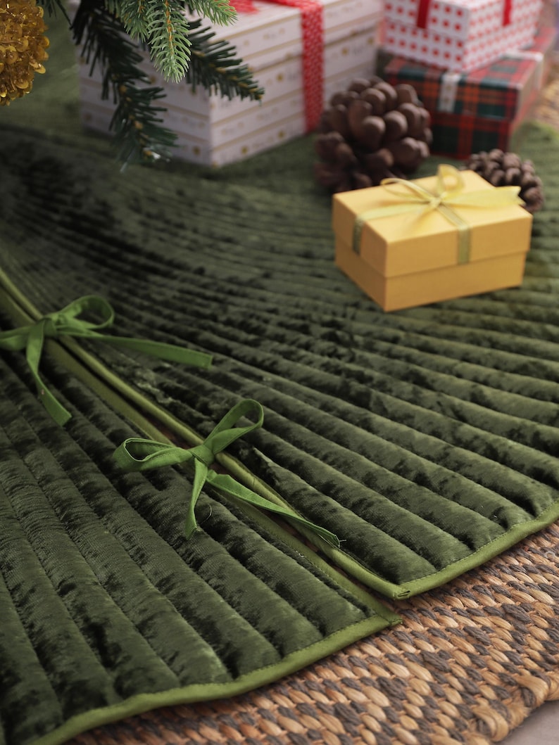 Puede incluir: Una falda de &aacute;rbol de Navidad de terciopelo plisado verde oscuro con un lazo verde. La falda est&aacute; sobre una estera tejida, con regalos envueltos y pi&ntilde;as en el fondo. Tambi&eacute;n se ve una peque&ntilde;a caja de regalo amarilla.