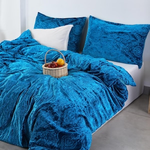 May include: A bed is dressed with a teal velvet duvet cover and matching pillowcases. A white pillow is placed on top. A small woven basket with fruit sits on the duvet. The bedding has a paisley pattern.