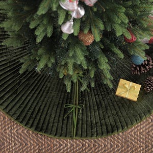 Puede incluir: Una falda de &aacute;rbol de Navidad de terciopelo verde con un dise&ntilde;o plisado. La falda rodea un &aacute;rbol de Navidad decorado con adornos y regalos envueltos. La falda est&aacute; sobre una superficie tejida marr&oacute;n.
