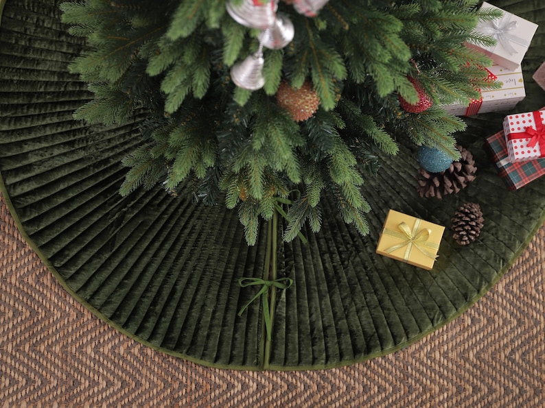 Puede incluir: Una falda de &aacute;rbol de Navidad de terciopelo verde con un dise&ntilde;o plisado. La falda rodea un &aacute;rbol de Navidad decorado con adornos y regalos envueltos. La falda est&aacute; sobre una superficie tejida marr&oacute;n.