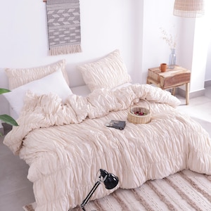 Pode incluir: Uma cama creme com um edredom e almofadas texturizadas. Uma pequena cesta de vime com frutas está na cama, junto com um livro. Uma luminária de mesa preta e um tapete listrado estão em primeiro plano. Uma tapeçaria tecida e uma pequena mesa de cabeceira de madeira completam a cena.